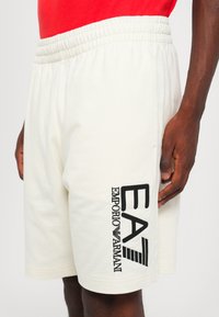 Herre shorts med elastisk linning i lys kremfarge, med en svart brodert "EA7 EMPORIO ARMANI" logo på siden.