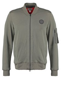 Giacca bomber verde oliva con zip frontale, polsini e orlo a costine, dotata di una toppa sul petto e di una tasca con zip sul braccio.