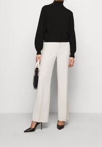 Svart höghalset tröja med ballongsärmar, kombinerad med vita vida byxor och svarta spetsiga klackar, hållandes en tvåfärgad väska.