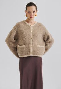 Beige fluffete cardigan med rund hals, gylne knapper og lommer foran, stylet over en brun satinlreport. Myk tekstur og løs passform.