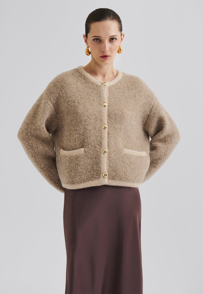 Beige fluffete cardigan med rund hals, gylne knapper og lommer foran, stylet over en brun satinlreport. Myk tekstur og løs passform.