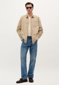 Beige cotton jacket with a chest pocket, layered over a light shirt, paired with blue denim jeans and dark shoes, glasses add contrast.