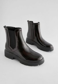 Botas negras de charol hasta el tobillo con paneles laterales elásticos, punta redondeada y suela de goma robusta. Textura suave con un diseño minimalista.