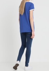 Camiseta de algodón azul con ribete blanco, diseño ajustado, combinada con jeans ajustados de mezclilla oscura y zapatillas blancas. La vista trasera muestra cabello largo.