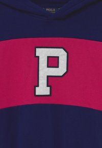 Námořnická modrá mikina s kapucí, která má vodorovný růžový pruh a velký bílý emblém "P" s černým obrysem na oblasti hrudi.