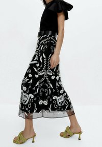 Jupe noire brodée avec des motifs floraux et abstraits blancs complexes, ourlet en transparence. Portée avec un haut noir et des chaussures tressées vertes.