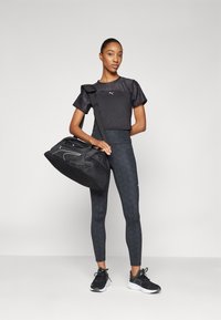 Top deportivo negro corto con logo, leggings negros con estampado y zapatillas negras. Sosteniendo un bolso de deporte negro con acentos en contraste y cremalleras.