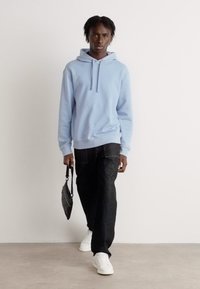 Sudadera con capucha de color azul claro, pantalones negros y zapatillas blancas. El modelo lleva una pequeña bolsa negra. Diseño minimalista, estilo casual.