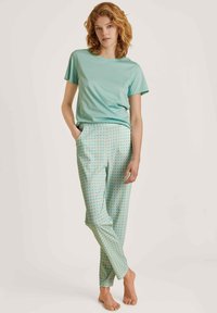 Lichtblauwe t-shirt gecombineerd met gemusterde, mintgroene broek met een abstract ontwerp. De broek heeft zijzakken; het model staat blootvoets.