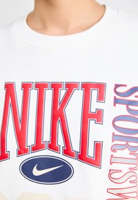 Balts džemperis ar spilgti sarkanu "NIKE" tekstu un zilu Nike logotipu. Audums izskatās gluds, ar atšķirīgām grafiskā dizaina elementiem.