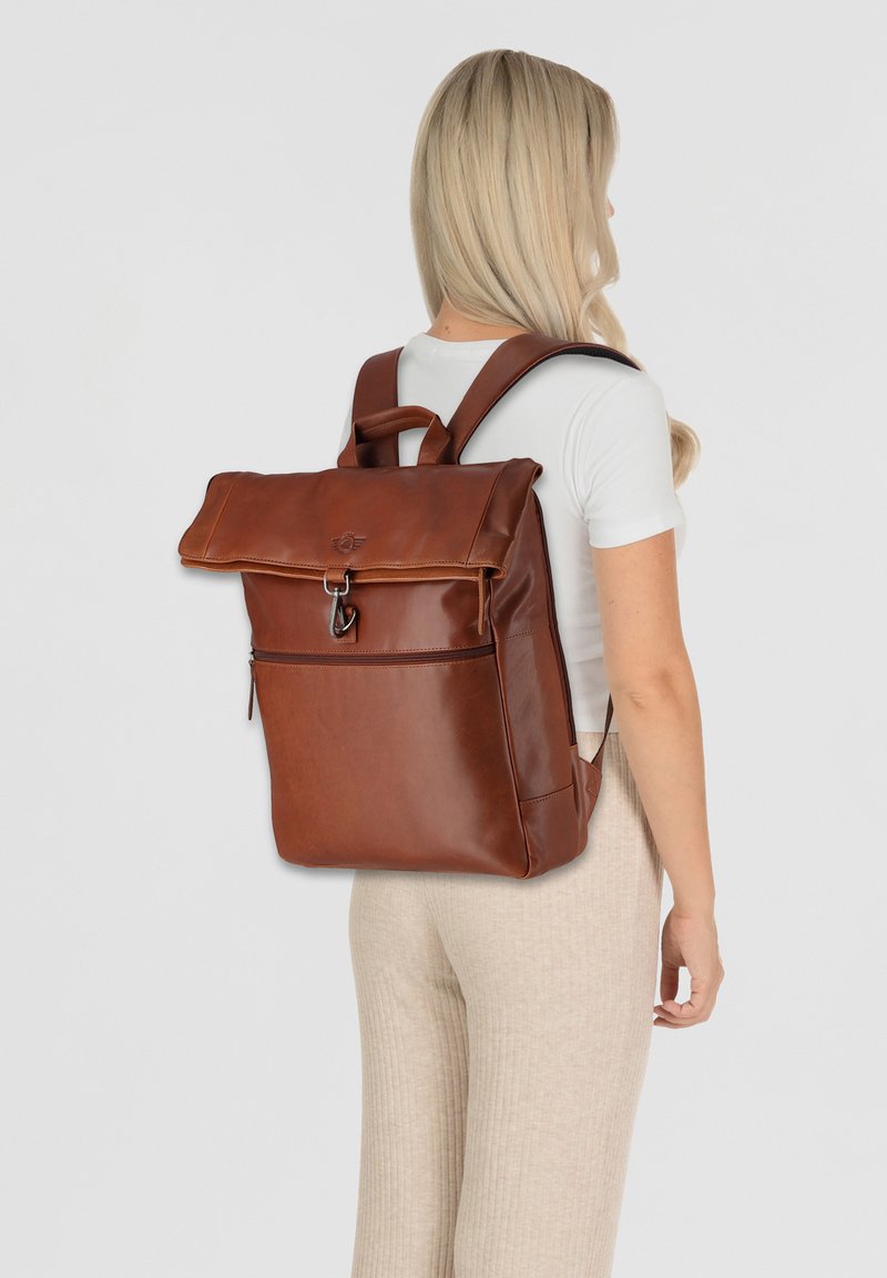 Sac à dos en cuir marron avec un rabat repliable, une poignée sur le dessus et des bretelles réglables. Comprend une poche zippée à l'avant et une texture lisse.