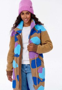 Uśmiechnięta kobieta w wielokolorowej polarowej kurtce z niebieskimi i fioletowymi łatami, różowej wełnianej czapce, białej koszuli i jasnoniebieskich dżinsach.