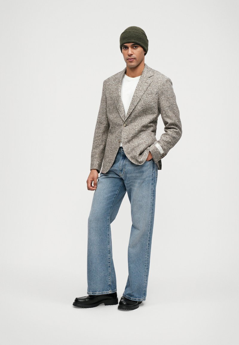 Grijze tweed blazer, wit T-shirt, lichtblauwe wijde jeans, zwarte schoenen, groene beanie. Blazer heeft gelapelle kraag en een enkele knoop.