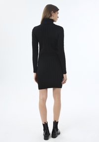 Frau steht mit dem Rücken zur Kamera in einem schwarzen Pulloverkleid mit Zopfmuster und schwarzen ankle boots vor einem schlichten weißen Hintergrund.