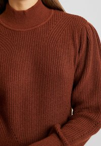 Bronzen ribgebreide trui met een hoge hals, voorzien van diagonale textuurdetails en gepofte mouwen. Gemaakt van een gezellig gebreid materiaal.