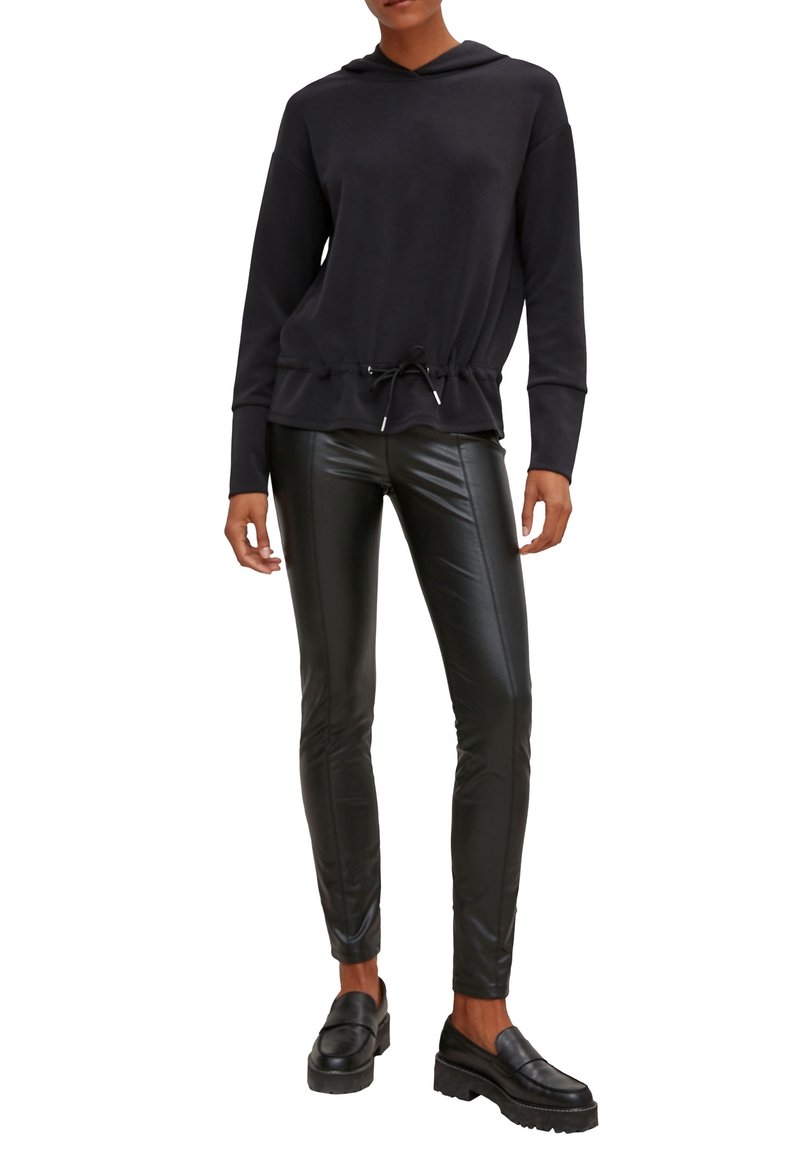 Femme portant un sweat à capuche noir avec cordon à la taille, pantalon noir en cuir synthétique et mocassins à plateforme noirs, debout sur fond blanc.