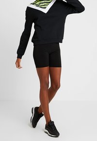Persona con pantalones cortos deportivos negros, zapatillas negras y blancas, y una sudadera negra con un diseño verde y blanco en el pecho.