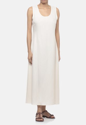 Vestito bianco semplice senza maniche lungo fino alla caviglia, indossato con sandali piatti marroni borchiati, in piedi su sfondo bianco.