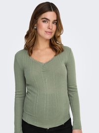 Grøn ribstrikket langærmet top med V-udskæring og bølget kant. Har lodret tekstur og centrale knapdetaljer. Kombineret med sorte bukser.