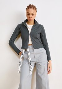 Veste zippée grise avec des manches longues, au design court, en tissu lisse ; associée à un pantalon large gris clair et à une écharpe blanche à motifs.