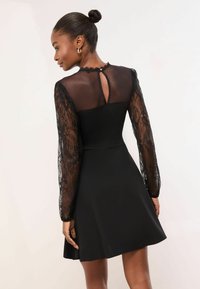 Rochie neagră cu mâneci lungi din dantelă, corset strâmt și fustă evazată; panou transparent și detaliu cu nasturi în spate. Accente de dantelă texturată pe întreaga rochie.