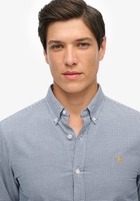 Camisa de cuadros en azul y blanco, de botones y con cuello, que presenta un logo de la marca en el lado izquierdo del pecho. Material de algodón, textura suave.