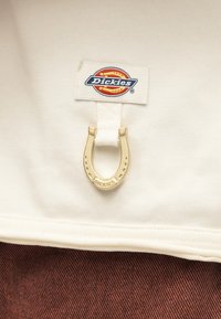 Υφάσμα κρεμ με ραμμένη ετικέτα της μάρκας Dickies και προσαρτημένο μικρό χρυσό γούρι σε σχήμα πεταλούδας πάνω σε καφέ υφή υφάσματος.