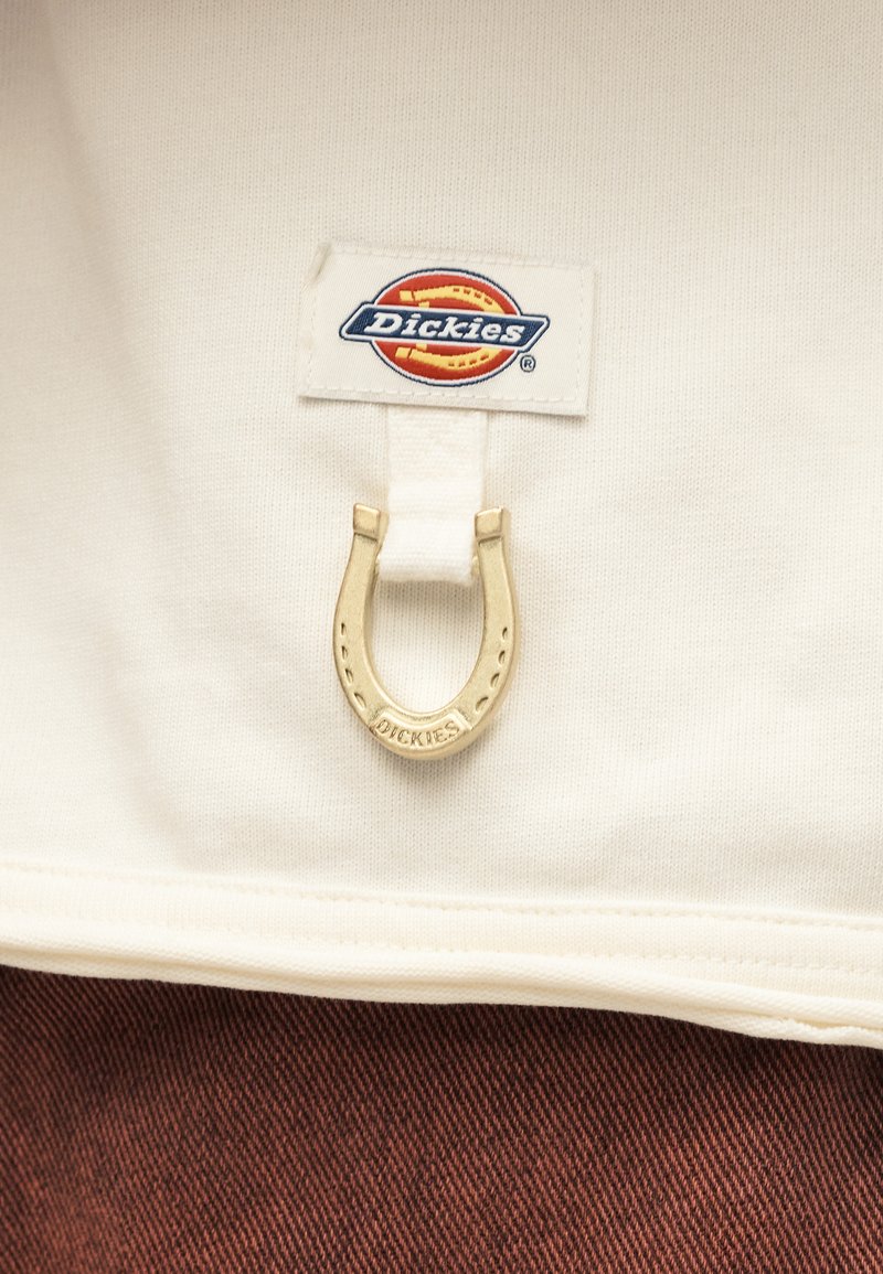 Cremiger Stoff mit einem aufgenähten Dickies-Markenlabel und einem kleinen goldenen Hufeisenanhänger darüber, auf braunem, strukturiertem Stoff.