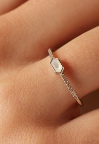 Rosenvergoldeter Ring mit einem hexagonalen weißen Steinzentrum und einem Band, das mit kleinen klaren Edelsteinen geschmückt ist, das auf der Haut einer Hand ruht.