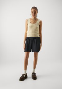 Knit cream tank top with scalloped edges, paired with black textured shorts. Model wears brown platform shoes and cream socks.