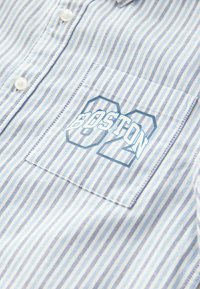 Gombos ing, vékony kék és fehér függőleges csíkokkal, bal oldali zsebbe helyezve, és kék "BOSTON" grafikával, vastag betűtípussal.