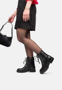 Botas de cuero negras hasta el tobillo con un acabado texturizado, cordones en la parte frontal, detalles en dorado y una suela resistente, combinadas con una falda negra.