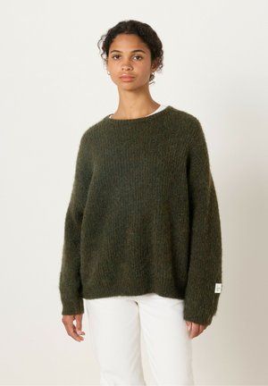 Donkergroene gebreide trui met een losse pasvorm, geribbelde textuur en brede mouwen. Met een klein label aan de linkerkant. Gedragen over witte broek.