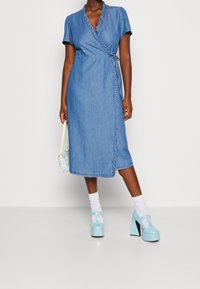 Denim omslagsklänning med korta ärmar, volangkant längs halsringningen; matchad med ljusblå klumpiga klackar och vita strumpor.