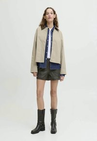 Beige kurz geschnittene Jacke über einem blauen Jeanshemd, einem weißen T-Shirt und dunklen grünen Shorts. Schwarze kniehohe Stiefel mit Schnallen runden das Outfit ab.