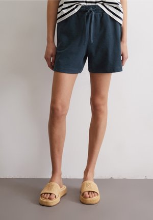 Personne portant un short bleu marine foncé à cordon, une chemise rayée et des sandales tissées beige, debout sur un sol gris contre un mur blanc.