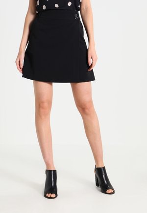 Kvinde iført en sort A-formet mininederdel med to knapper i taljen, sorte åbne ankelstøvler med hæl og en sort top med polkaprikker.