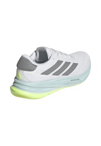 Zapatilla deportiva blanca con acentos grises, parte superior de malla texturizada, suela acolchada en azul claro y elevación en amarillo neón, con el logo de las tres rayas.