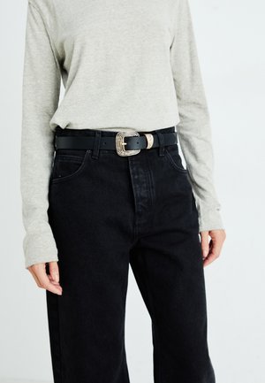Black leather belt with a square, textured gold buckle worn over black jeans and a light gray long-sleeve shirt. Simple design, modern look.