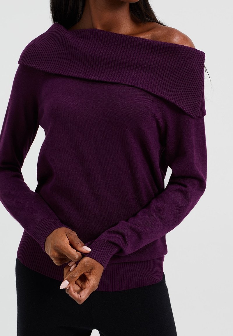 Femme portant un pull violet à manches longues, épaules dénudées, les mains jointes devant un fond uni.