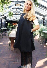 Fekete ujjatlan ruha, laza szabással és mintás csipke alsó résszel. A modell hullámos szőke hajjal rendelkezik, és ezüst órát visel. Zöld háttér.