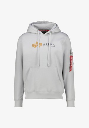Jasnoszara bluza z kapturem Alpha Industries z przednią kieszenią, kapturem z regulowanym sznurkiem, logo w kolorze pomarańczowo-czarnym oraz kieszenią na rękawie zamykaną na zamek z czerwoną metką "Remove Before Flight".