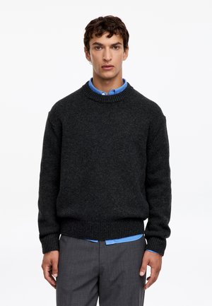 Jeune homme portant un pull en maille gris foncé par-dessus une chemise bleue à col et un pantalon gris, debout devant un fond blanc uni.