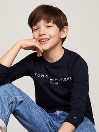 Mörkblå skjorta med långa ärmar och vit text "TOMMY HILFIGER EST. 1985". Denimjeans med ljus tvätt och uppvikta ärmar. Leende pojke som vilar hakan på handen.