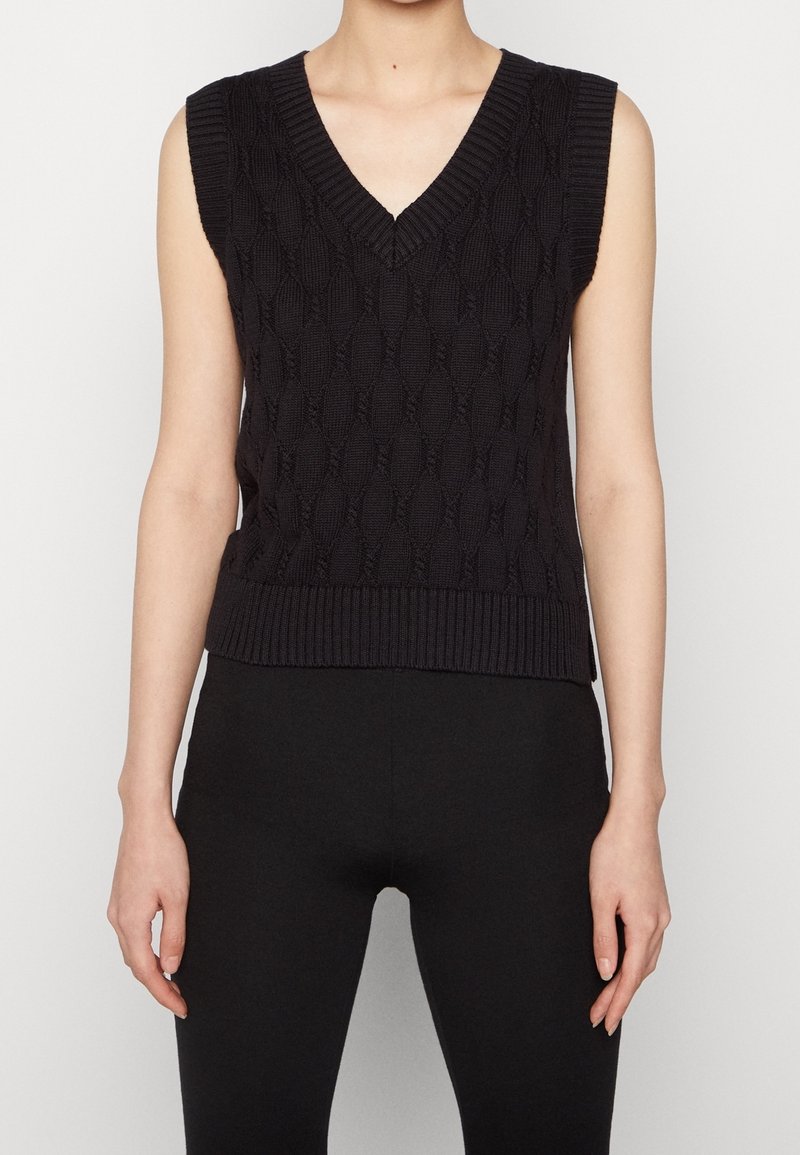 Svart ärmlös stickad väst med V-hals, prydd med ett diamantmönster och ribbad nederkant. Bärs över svarta leggings.