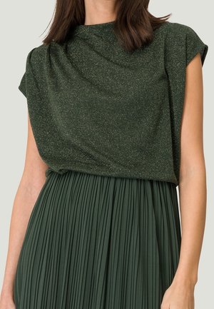 Woman wearing a dark green glittery sleeveless top paired with a matching dark green pleated skirt.