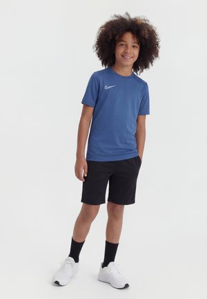 Enfant souriant aux cheveux bouclés portant un T-shirt Nike bleu, un short noir, des chaussettes noires et des baskets blanches, debout devant un fond uni.