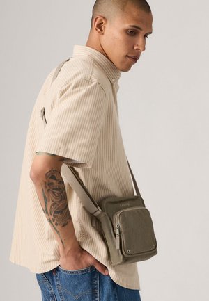 Man wearing beige striped shirt and blue jeans with an olive-green Levi's crossbody bag hanging by his side and tattooed forearm.