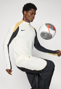 Nike majica dugih rukava u krem boji s crnim i zlatnim detaljima, uparena s tamnim hlačama. Igrač balansira nogometnu loptu na koljenu.