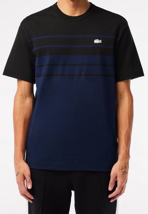 Homme portant un t-shirt à manches courtes rayé bleu marine et noir avec un petit logo Crocodile blanc sur la poitrine, debout les bras détendus le long du corps.